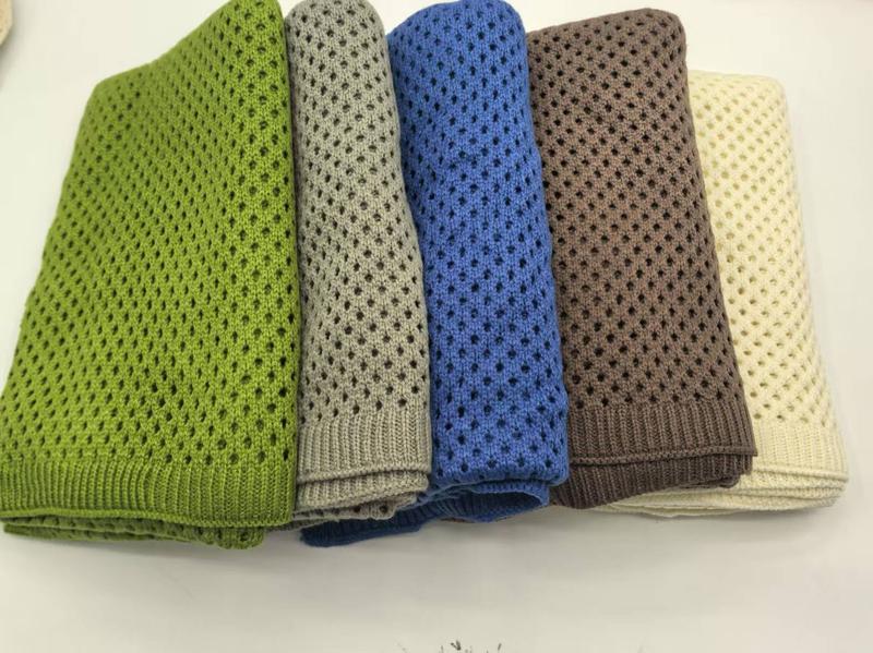 Five folded open-weave knit cotton throw blankets in green, grey, blue, brown, and cream stacked on white surface
