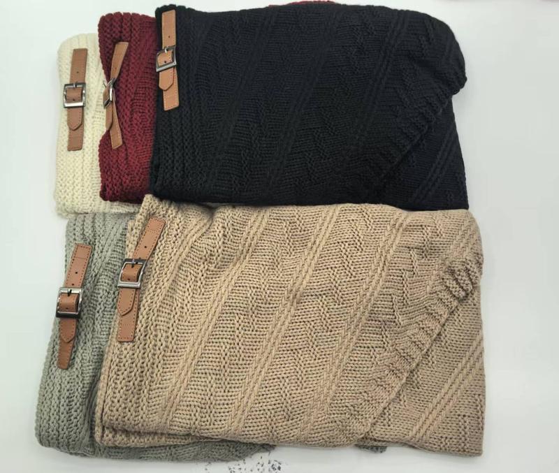 Five folded chunky cable knit throw blankets in beige, grey, black, burgundy, and cream with brown leather buckle straps