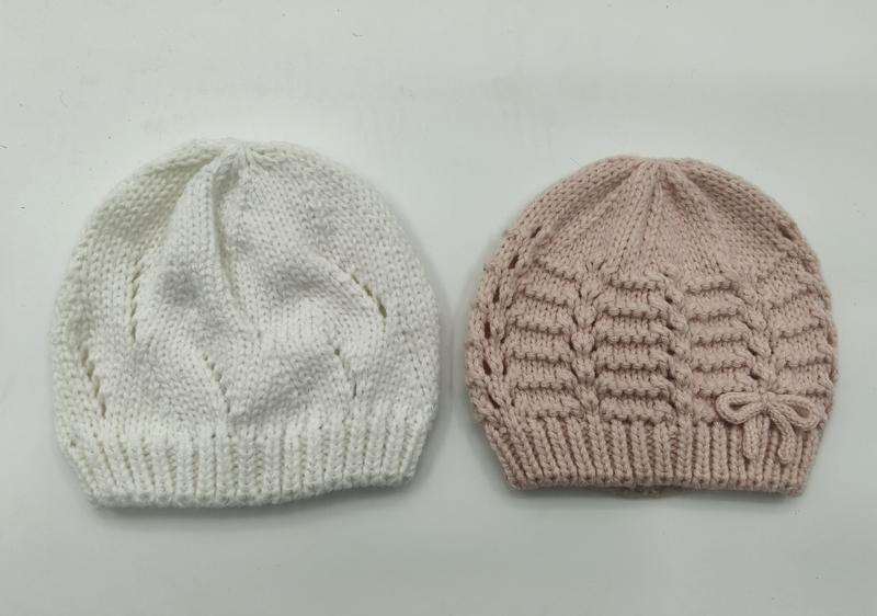 Two hand-knitted baby beanies side by side, white with leaf pattern and blush pink with horizontal texture and bow detail