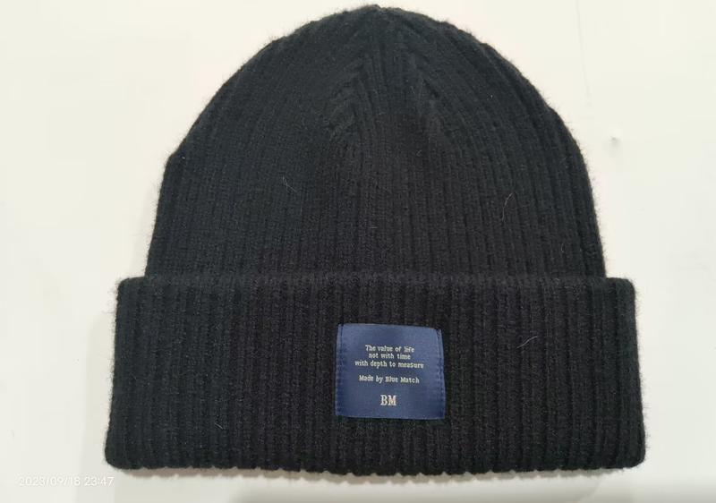Black ribbed knit cuffed beanie with blue woven label patch on folded brim, soft textured wool winter hat