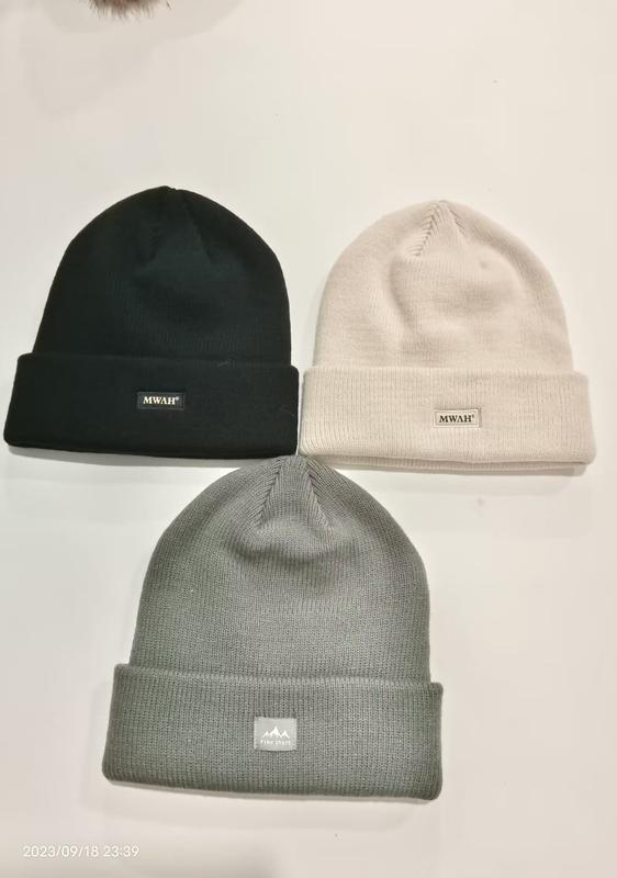 Three ribbed knit cuffed beanies laid flat in black, cream, and grey with small brand patch labels on white background