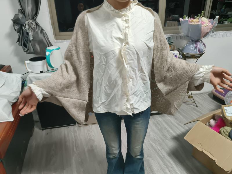 Woman wearing oversized beige knit cardigan layered over cream quilted ruffle-collar shirt with blue flare jeans indoors