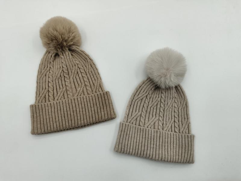 Two cable knit winter beanies with faux fur pom poms in taupe and light grey, displayed on white background
