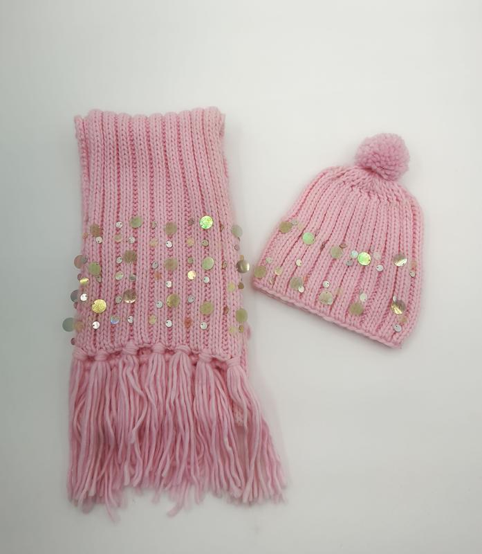 Pink ribbed knit winter scarf with fringe and sequins paired with matching pom-pom beanie hat on white background