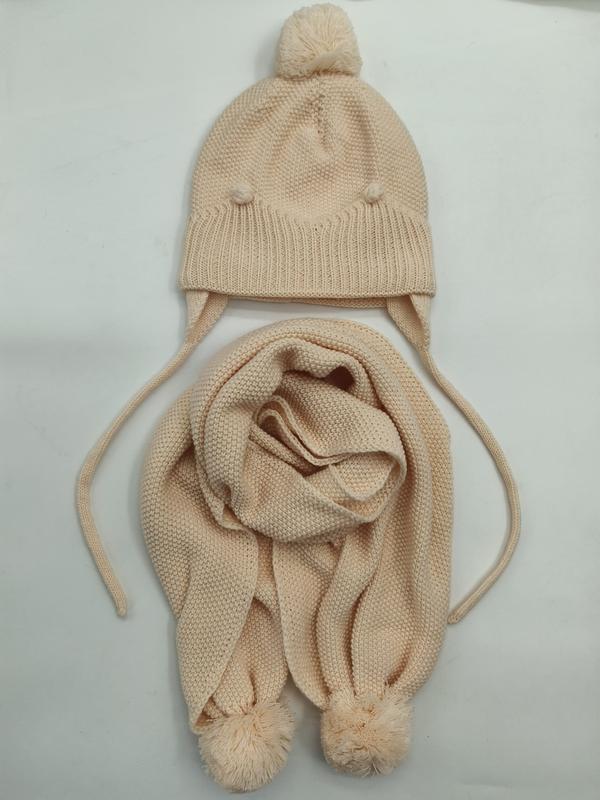 Cream knit children's winter hat with pom-pom top and ear flaps paired with matching knit scarf with pom-pom ends