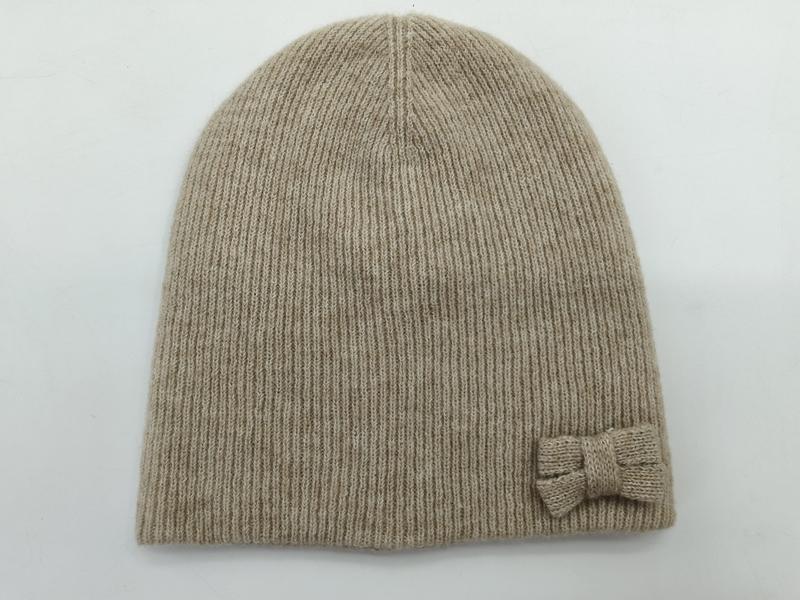 Beige ribbed knit beanie hat with small knitted bow accent on the side, laid flat on white background