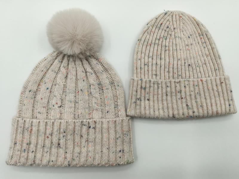Two cream speckled knit winter beanies side by side, one cable-knit with faux fur pom-pom, one ribbed with folded cuff