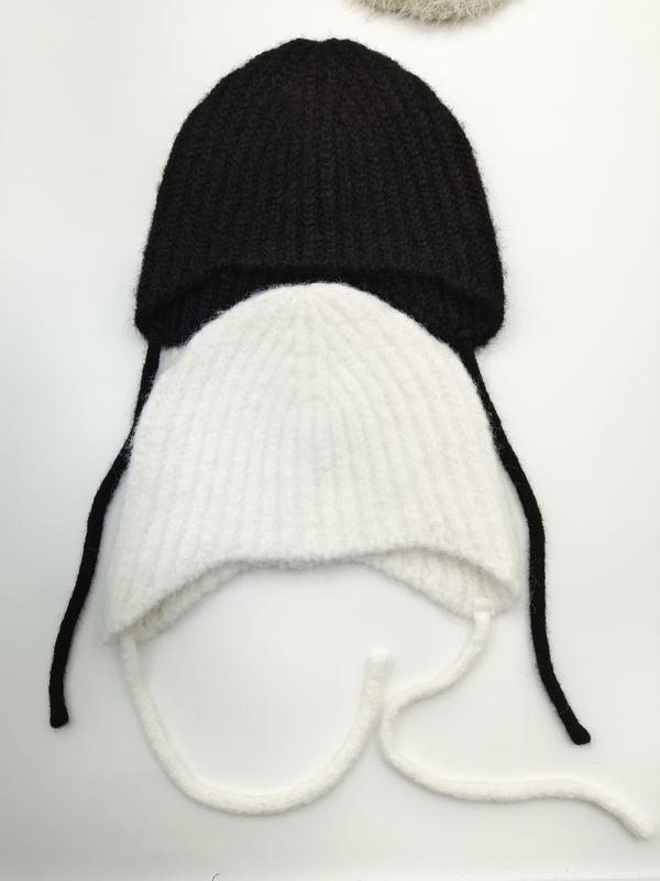 Two ribbed knit baby earflap hats with chin ties laid flat, one black and one white, on white background