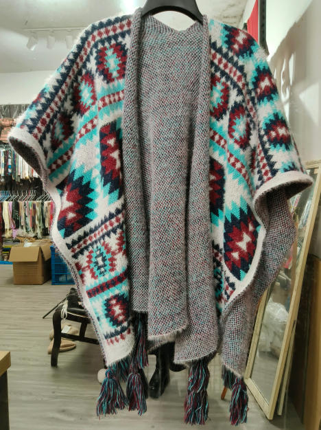 Open-front knitted poncho with Aztec geometric pattern in teal, burgundy and white, featuring tassel fringe hem, displayed on hanger