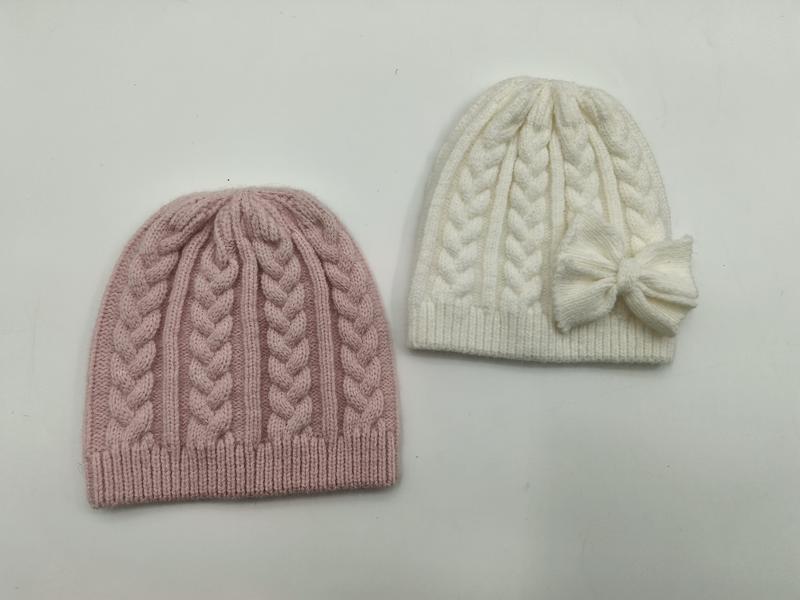 Two cable knit winter beanies side by side, one dusty pink and one cream white with a decorative bow on the side
