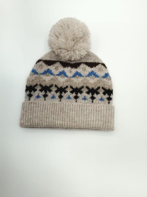Oatmeal knit beanie hat with Fair Isle snowflake pattern in blue and black, topped with a fluffy cream pom-pom